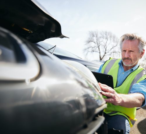 Mature Male Loss Adjuster With Digital Tablet Assessing Insurance Claim In Car Pound